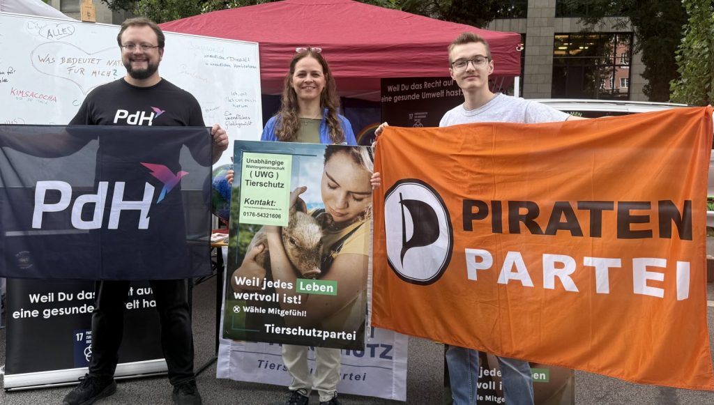 Drei Menschen stehen nebeneinander, lächeln und zwei zeigen die Flagge ein Plakat ihrer Partei. v.l.n.r. PdH, Tierschutz, PIRATENPARTEI